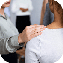 A person gently places a comforting hand on another person's shoulder, offering support. Other individuals are visible in the blurred background, suggesting a group setting.