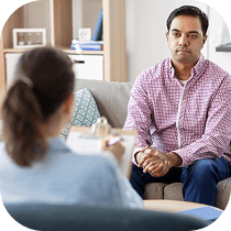A man in a pink checkered shirt sits on a couch, facing a woman with a clipboard, suggesting a therapy or counseling session in a comfortable office setting.