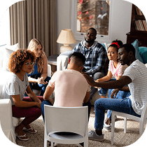 A diverse group of six people sit in a circle in a cozy living room, engaged in a supportive group discussion. One person has a comforting hand on another’s shoulder.
