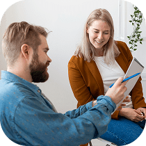 A man and a woman are sitting and smiling as the man shows something to the woman on a tablet. The woman is wearing a brown jacket and the man is wearing a blue shirt.