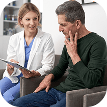 A healthcare professional shows a digital tablet to a seated man who touches his jaw, appearing to discuss a health concern or receive medical advice in a clinic setting.