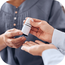 Close-up of one person handing a small white medication bottle to another person, who is holding out their hand. Both individuals are wearing long-sleeved shirts.