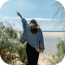 A person with long hair, wearing a denim jacket and black skirt, walks toward a sandy beach and the ocean, raising one arm while surrounded by greenery under a blue sky.