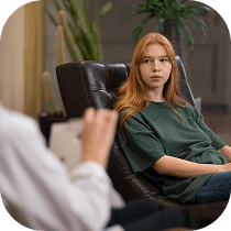 A young person with long red hair sits on a chair, looking at someone who appears to be a therapist taking notes. There is a plant in the background, and the setting looks like an office or counseling room.