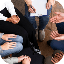 A small group of people sits in a circle, with their hands visible and one person holding a clipboard and pen, suggesting a meeting or support group session.