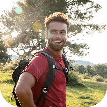 A smiling man with a beard and a backpack stands outdoors in a sunny, grassy area with trees in the background.
