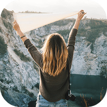 A person with long, blonde hair stands facing a rocky cliff and lake, holding up a white cloth above their head. The scene is outdoors, conveying a sense of freedom and adventure.