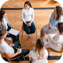 Five people sit in a circle on chairs in a bright room with wood floors, engaged in conversation. One person holds a clipboard, and another holds a cup.