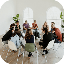 A diverse group of people sits in a circle of chairs in a bright room with large windows, engaged in a group discussion or support meeting.