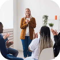 A woman stands smiling at the front of a bright room while four people seated around her applaud. There are plants and shelves in the background, and the group appears to be participating in a casual meeting or workshop.