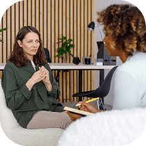 A woman sits and talks with a therapist who is taking notes in an office setting with a wooden wall and plants in the background.
