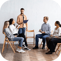 A group of five people sit in a circle on wooden chairs, attentively listening to a man standing and holding a clipboard, suggesting a meeting or group discussion in a bright room.