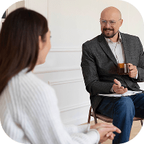 A man in a blazer holding a cup and a clipboard sits facing a woman in a white sweater, engaged in conversation in a bright, minimalist room.