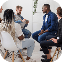 A group of five people sit in a circle on chairs, engaged in a friendly discussion in a bright room with white walls and plants.