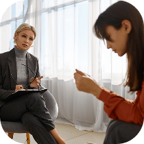 A therapist sits with a clipboard, attentively listening to a woman who looks down and holds something in her hands during a counseling session in a brightly lit room.