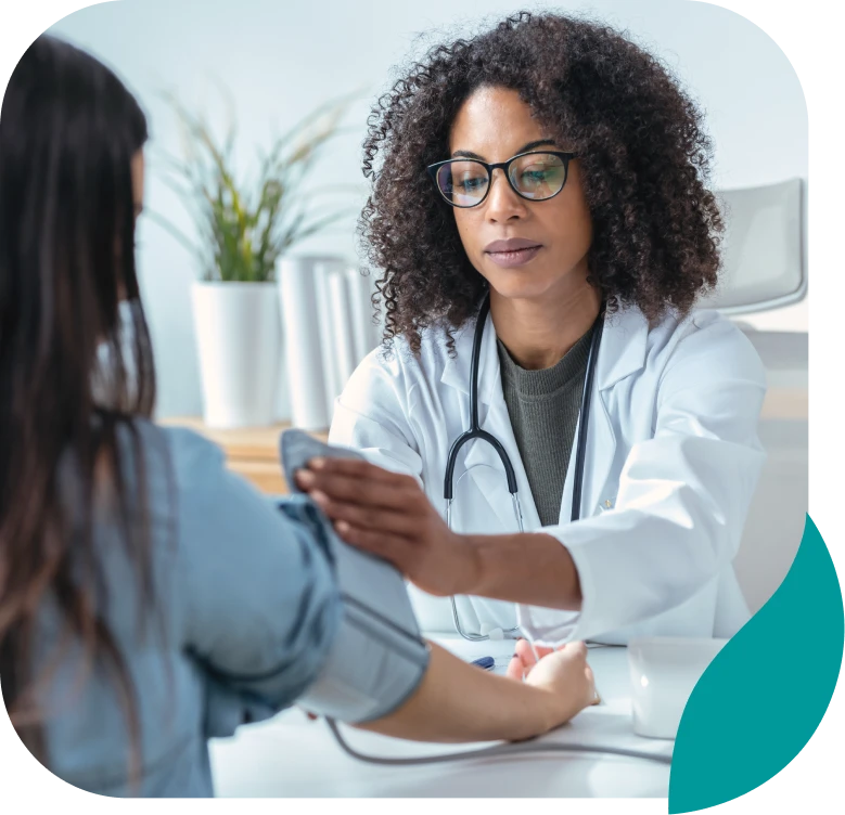 A female doctor with curly hair and glasses checks a patient’s blood pressure in a bright medical office, with a stethoscope around her neck and a plant in the background.