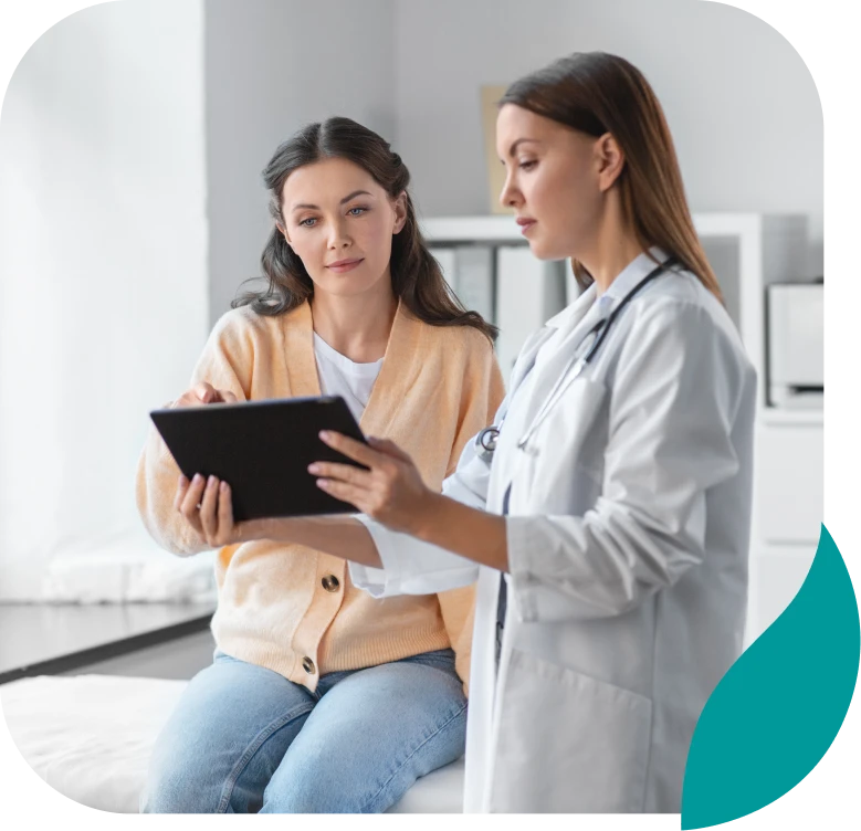 A female doctor in a white coat shows a tablet to a woman sitting on an exam table. The woman listens attentively, and they appear to be discussing medical information in a bright, modern office.
