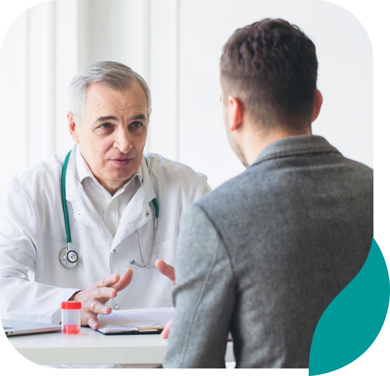 A doctor in a white coat with a stethoscope talks to a male patient across a desk in a bright office. A small medical sample container is on the desk.