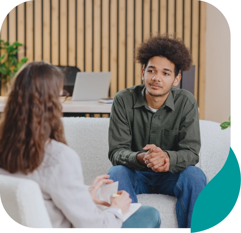 A young man with curly hair sits on a white couch, attentively listening to a woman with long brown hair across from him. They appear to be having a serious conversation in a modern, well-lit office space.